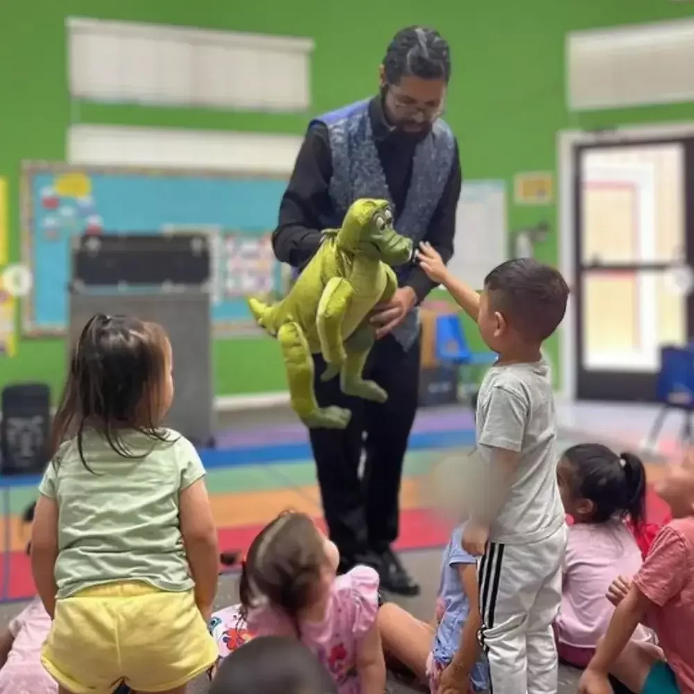 Preschooler reaching out to touch a dinosaur puppet during a classroom puppet show in Fremont, CA